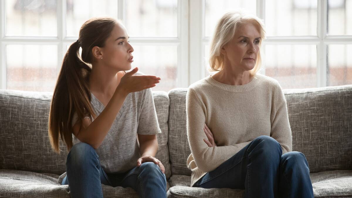 A mother and her adult daughter sitting side by side on a couch. The mother has her arms folded and is looking away while the daughter speaks to her critically.