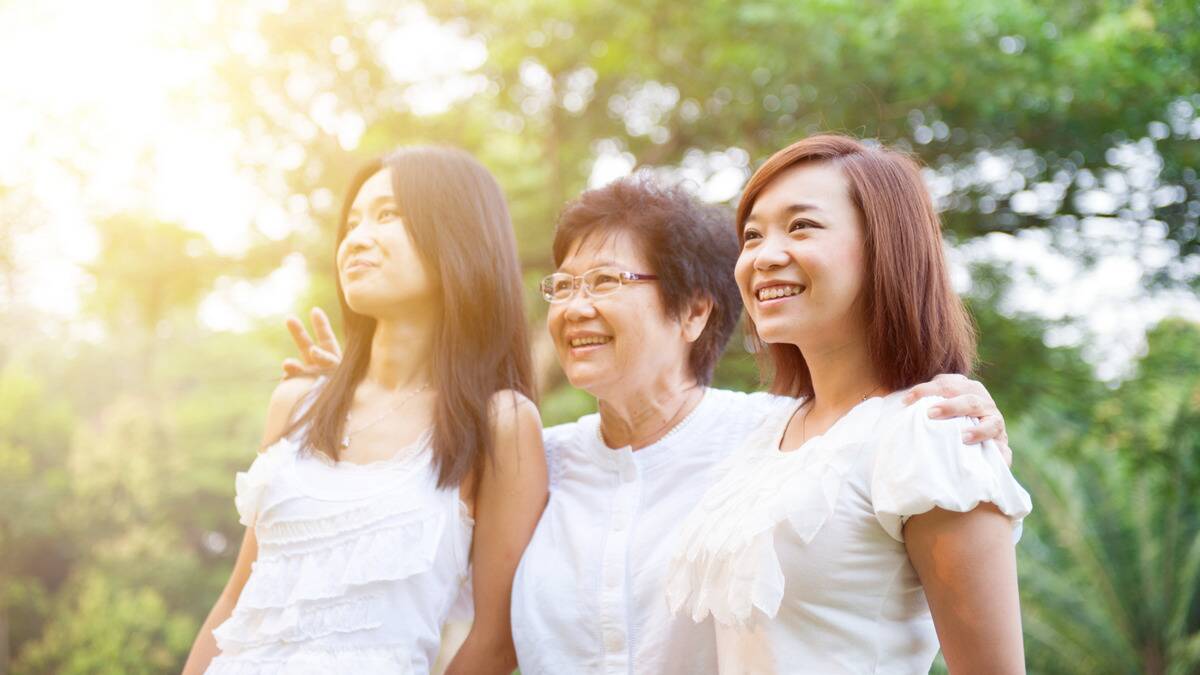 A mother standing outside with her two daughters, all smiling as they look forward, the mother's arms around them.