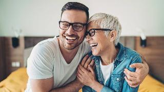 A mother and her adult son sitting side by side, both laughing, half-embracing one another.