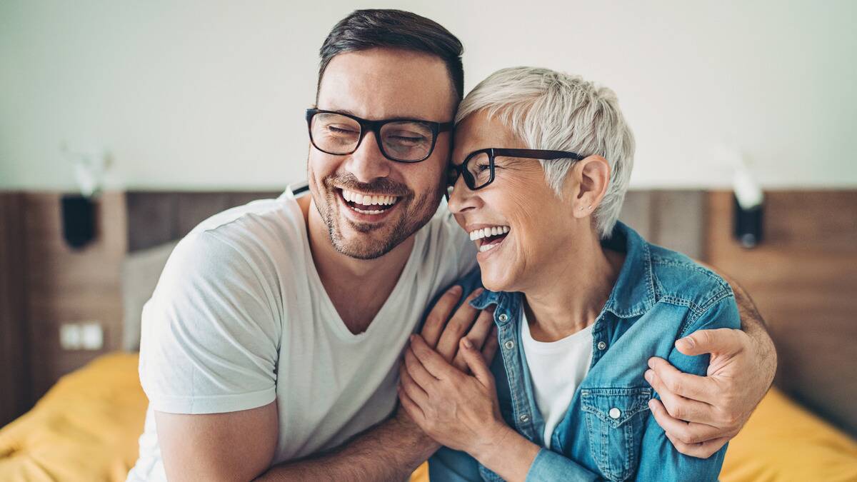 A mother and her adult son sitting side by side, both laughing, half-embracing one another.