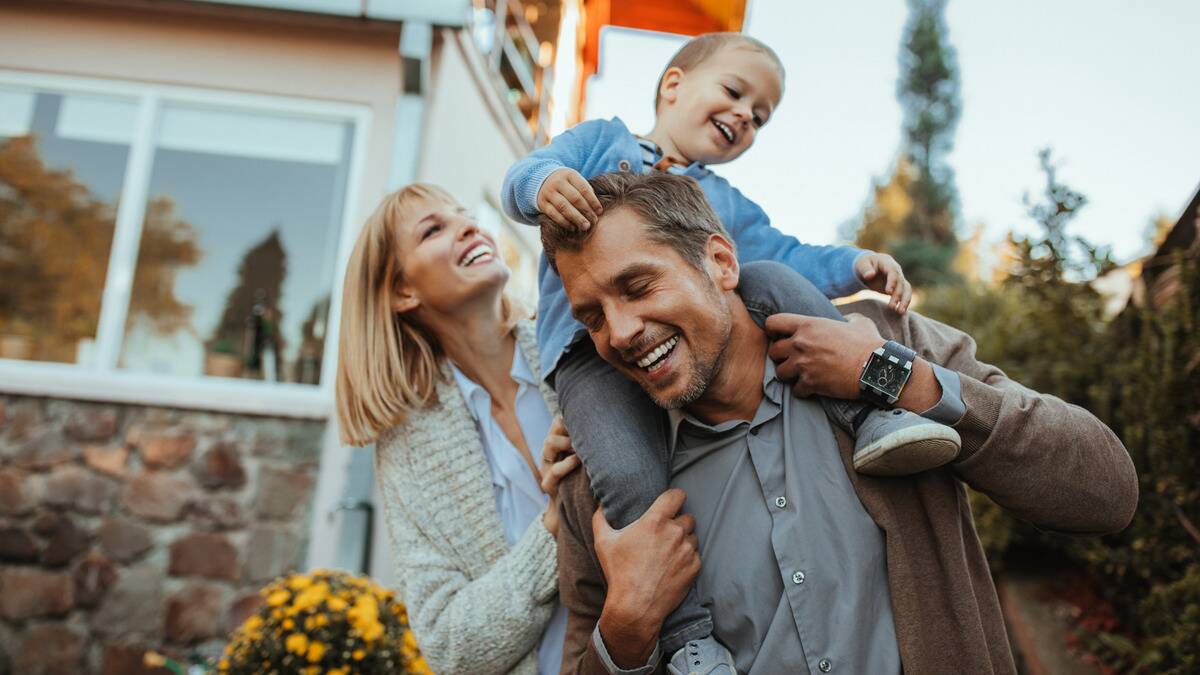 A mother and father playing with their young son outside, the son on his father's shoulders.