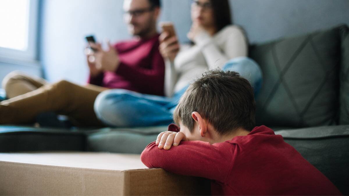 A young boy leaning against a table, hiding his face in his arms. In the background, both of his parents can be seen on their phone.