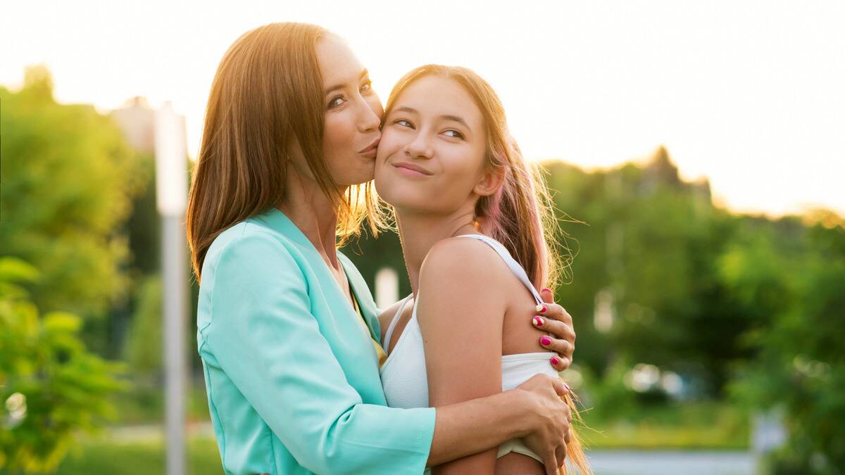 A mother kissing her teen daughter on the cheek, the daughter looking playfully annoyed.