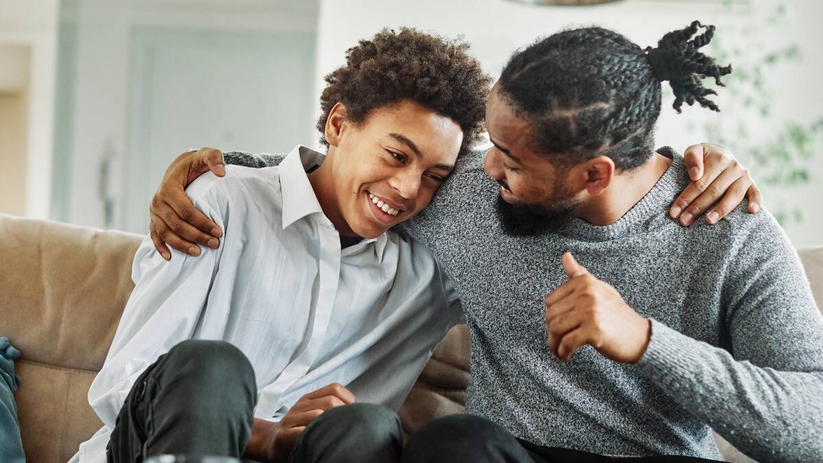 A father and son sitting side by side on a couch, embracing one another and smiling as they chat.