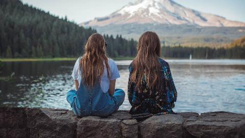 Two friends sitting outside in front of the water, facing away from the camera. 