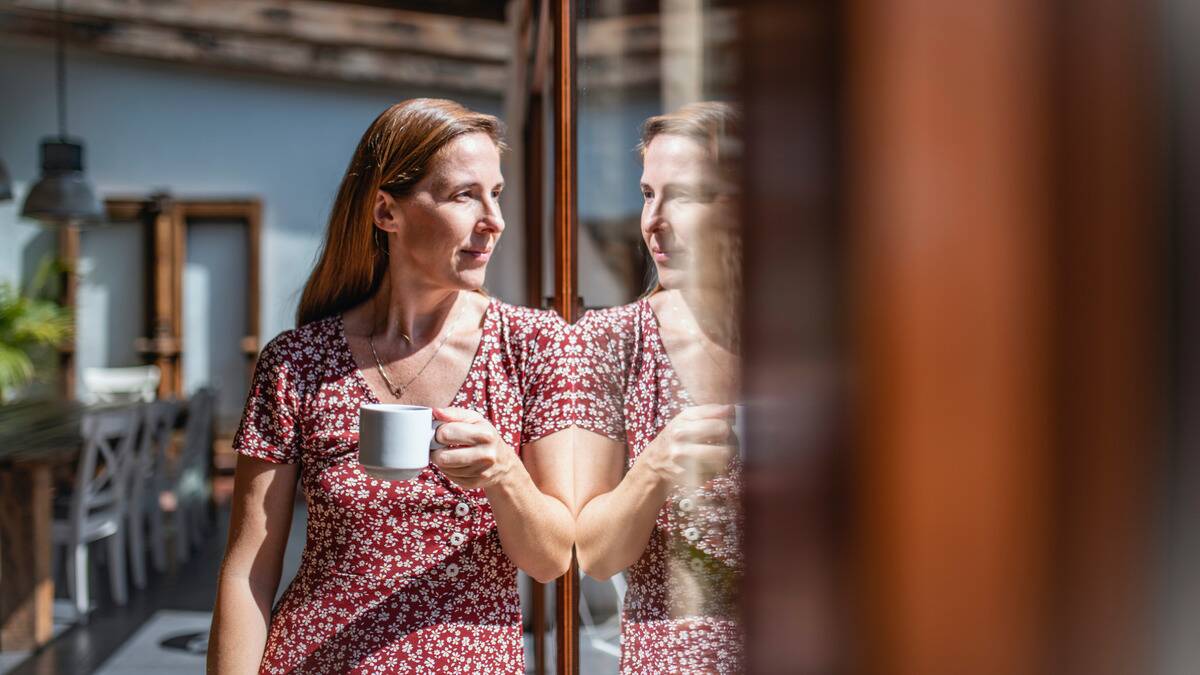 A woman holding a coffee mug and leaning against a windowed wall in which her reflection can be seen. 