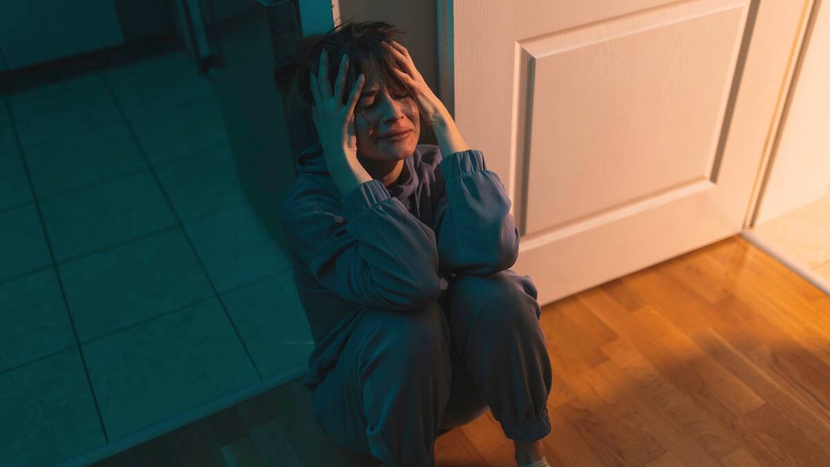 A woman sitting on the floor of her home, knees to her chest, hands on her head, visibly crying in the light that's coming in from the doorway.