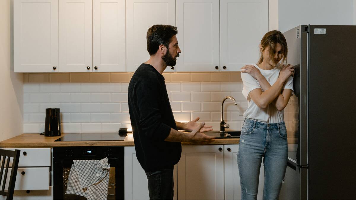 A woman hugging herself and leaning against the kitchen fridge while a man speaks sternly to her.