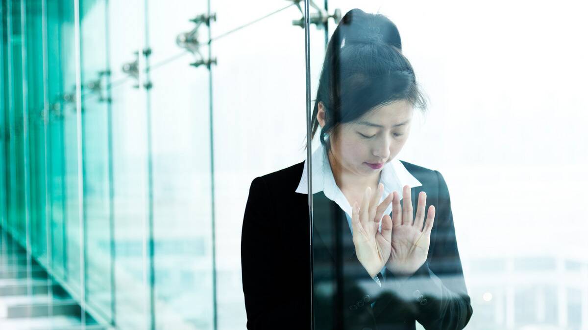 A business woman leaning against a glass wall, pushing her hands against it, looking down at herself dejectedly. 