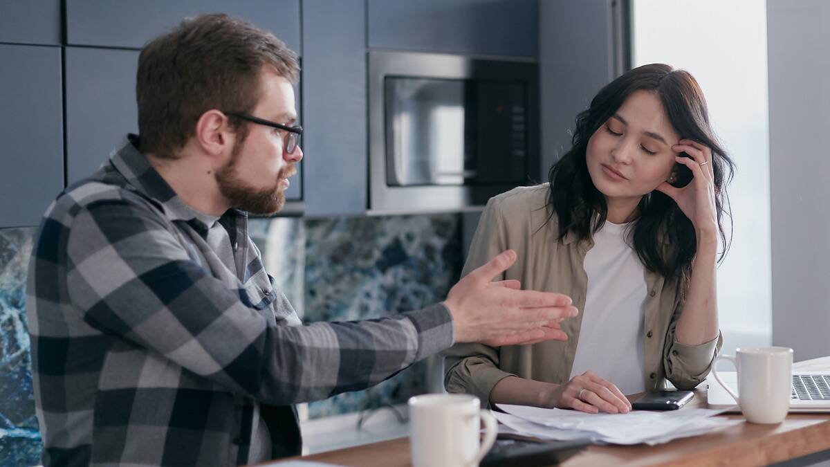 A woman looks stressed, eyes closed and resting her head in her hand, while a man sitting hear her speaks sternly to her.