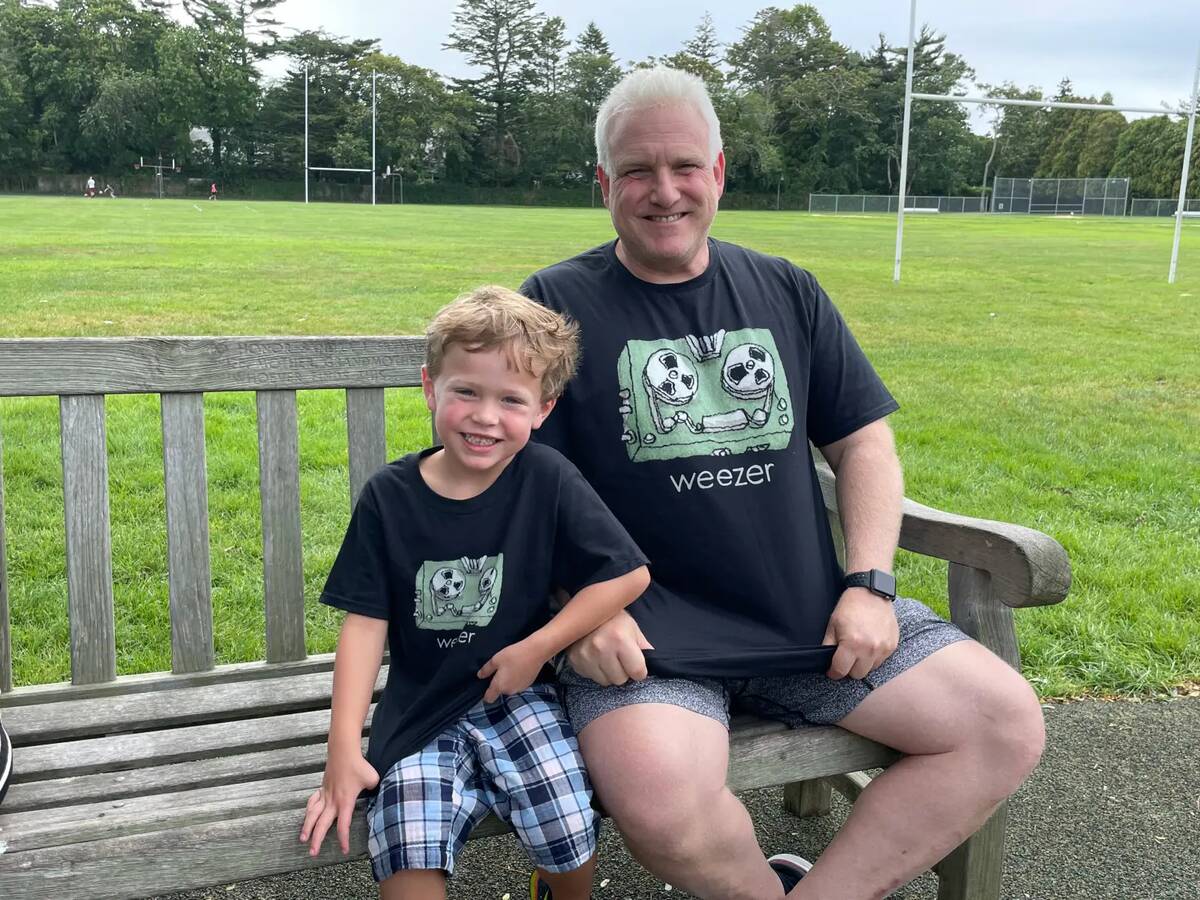 Stuart with his grandson on a park bench, both wearing matching Weezer shirts as they smile at the camera.
