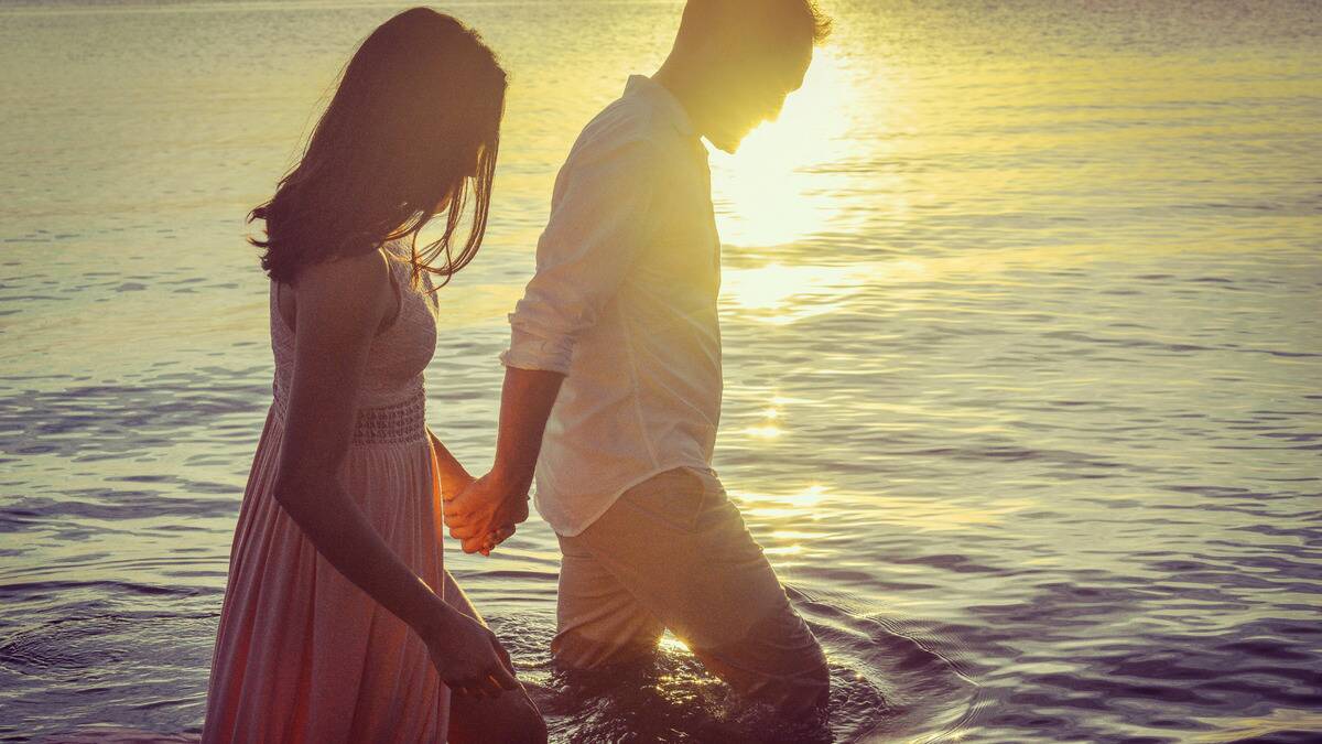 A couple walking through water at the beach, holding hands, the sun setting behind them.