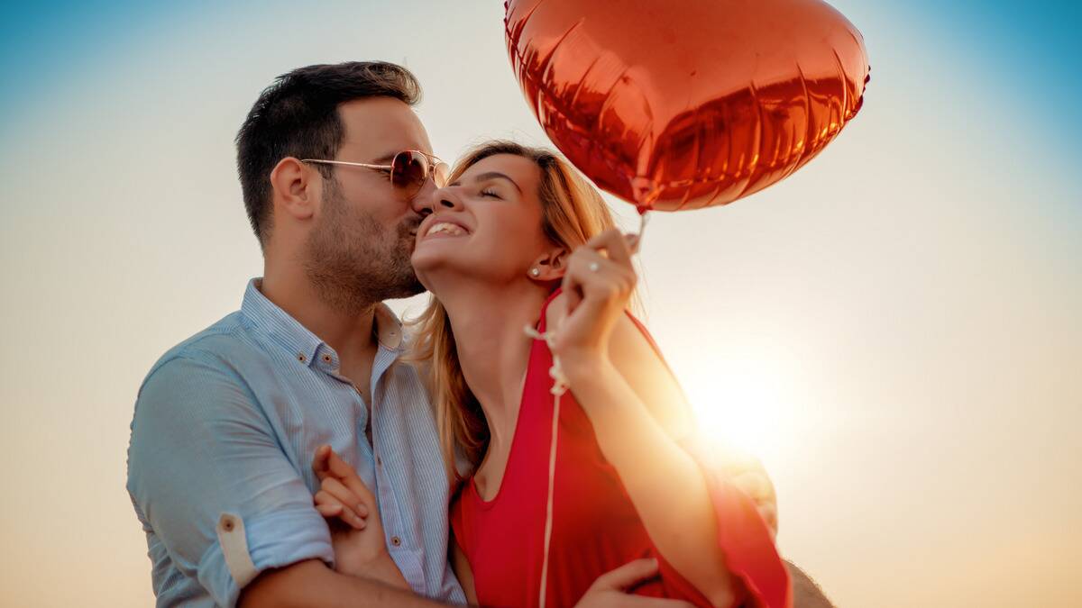 A couple standing with the sun behind them, the man kissing the woman's cheek, the woman leaning into him and holding a heart-shaped balloon. 