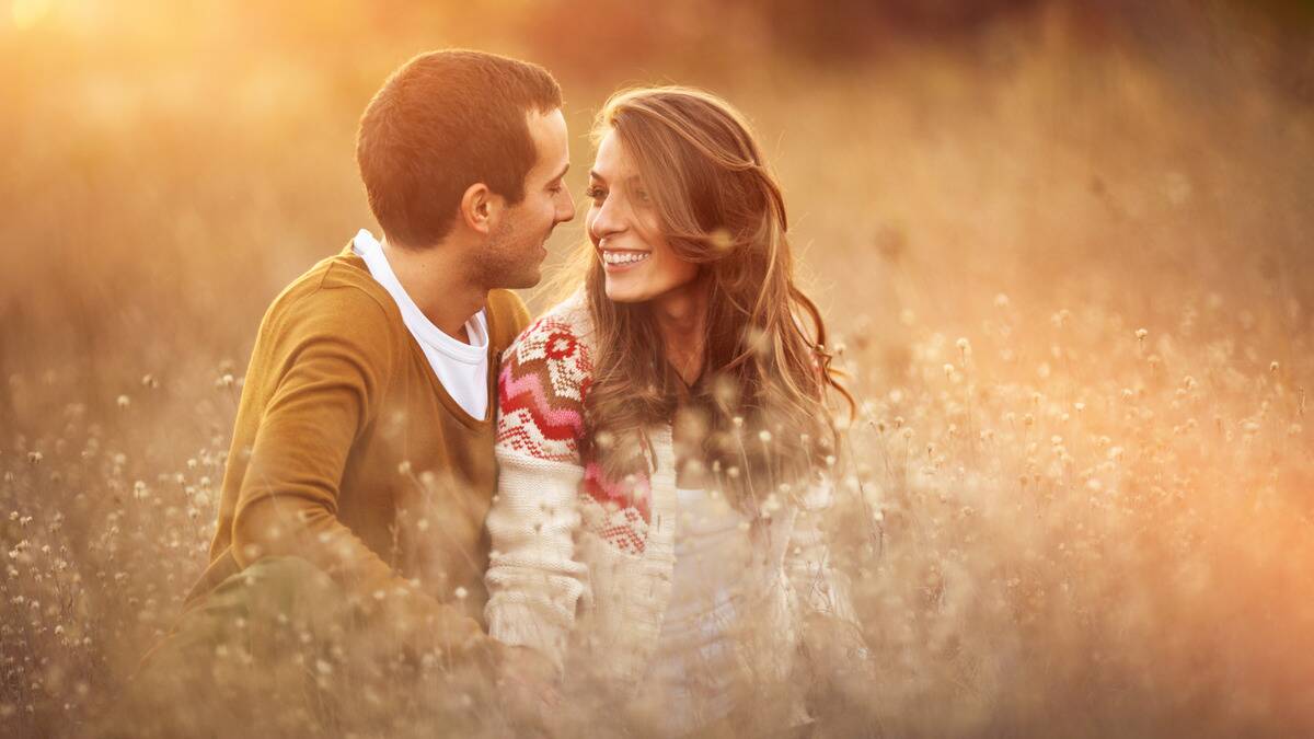 A couple sitting in a field of tall grass, smiling at one another.