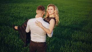 A man, faced away from the camera, carrying his girlfriend bridal style across a field as she smiles into the camera.