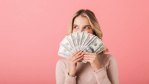 A woman smiling against a pink background, though her smile is hidden behind a fanned out stack of $100 bills.