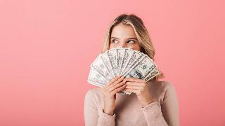 A woman smiling against a pink background, though her smile is hidden behind a fanned out stack of $100 bills.