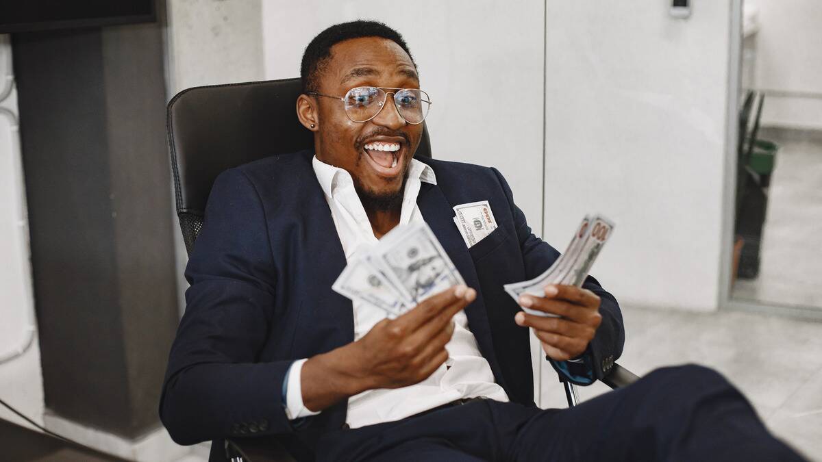 A man smiling excitedly as he holds a few $100 bills in both hands, more seen in his blazer breast pocket.