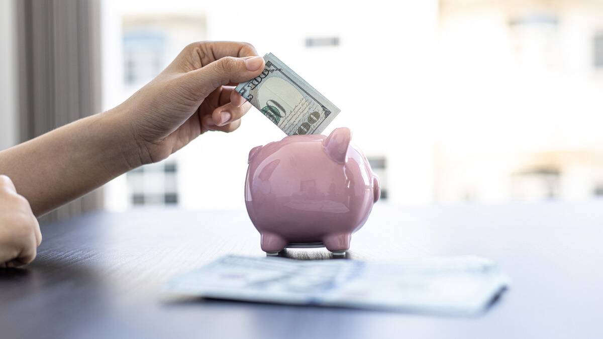 Someone putting a folded $100 bill in a small, pink, ceramic piggybank.