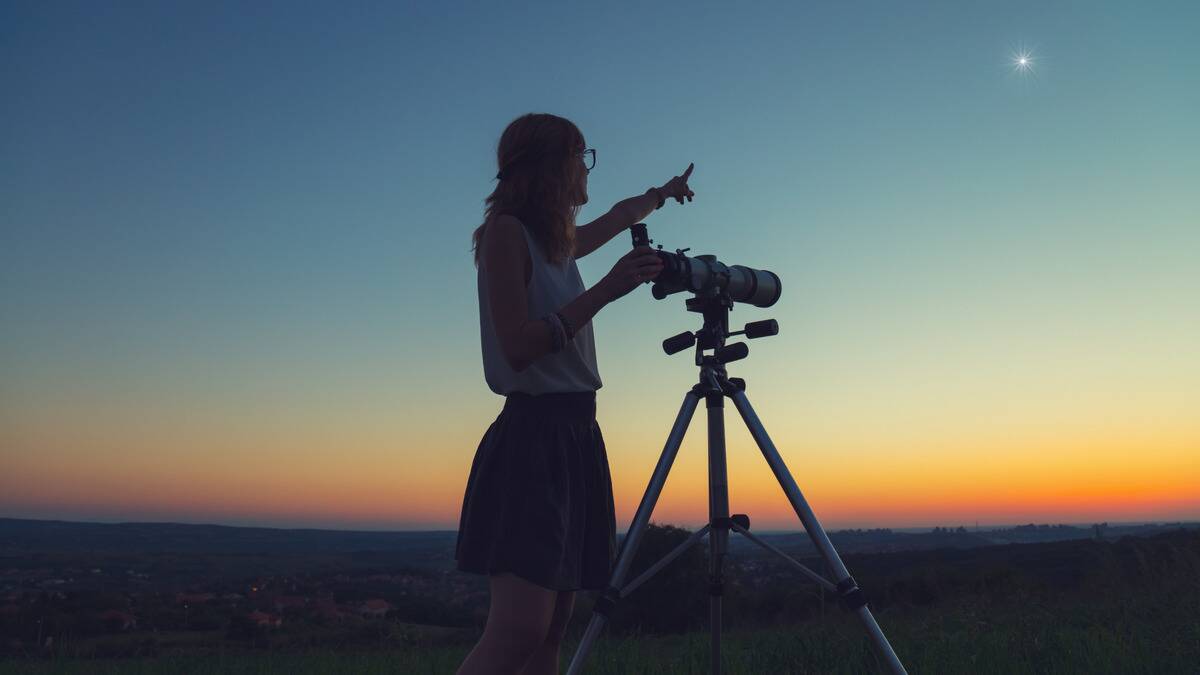 A woman standing at a telescope in the early morning, pointing toward one bright glowing point, probably a planet.