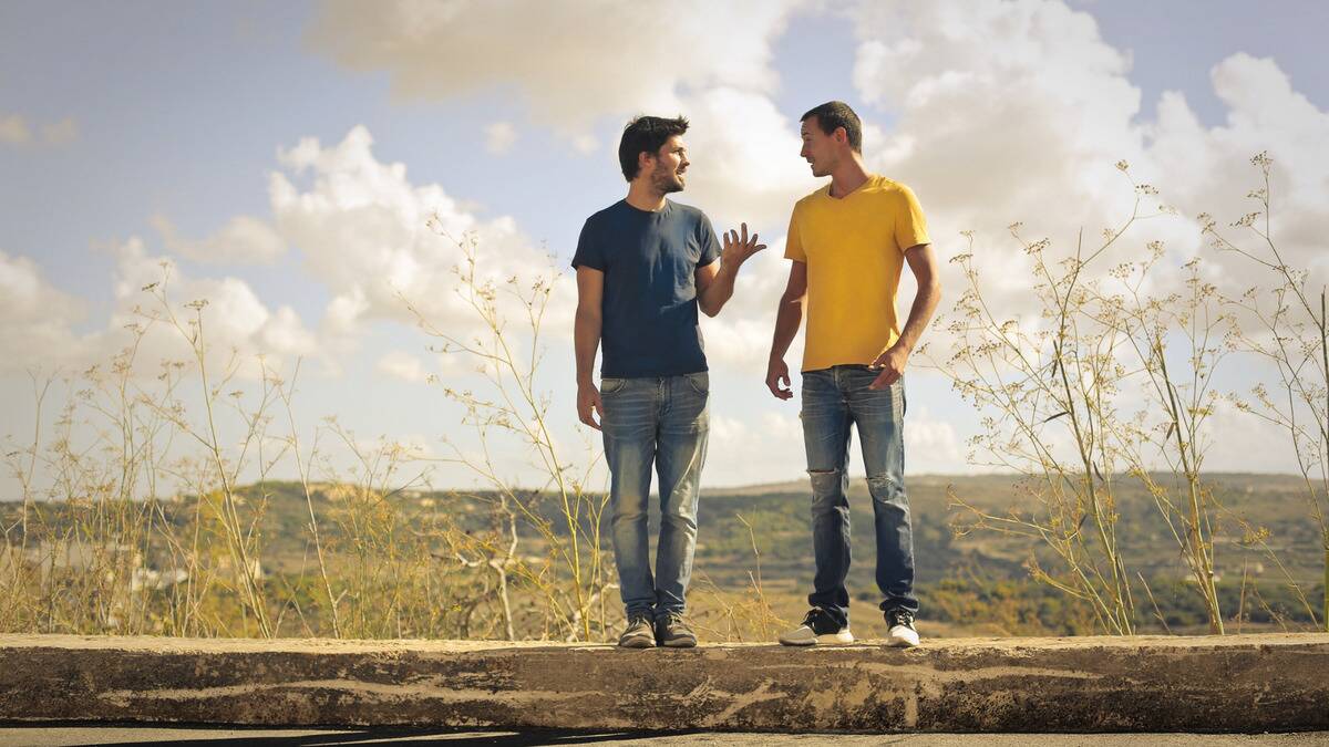Two friends standing side by side on a curb overlooking a landscape, chatting with each other.