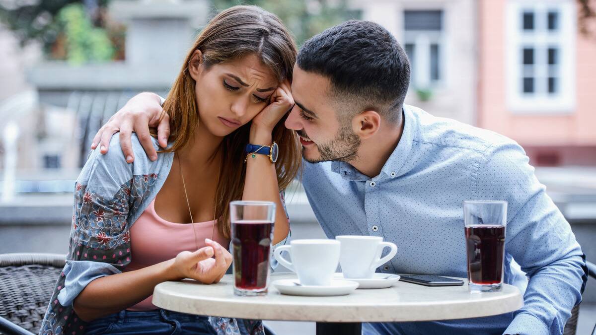 A couple sitting at a cafe table, the woman looking sad while the man wraps his arm around her and gets in her face with a smile.