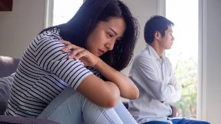 A couple sitting next to one another, the woman looking sad as she curls in on herself, the man with his arms crossed looking out the window.