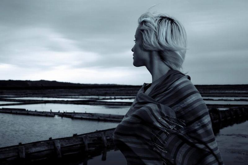 A greyscale image of a woman standing by the water, looking out into the distance.