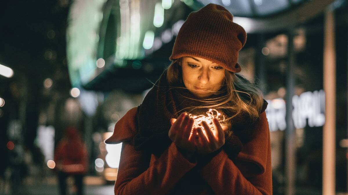 A woman walking outside in fall clothing, holding a bundle of fairy lights in cupped hands close to her face.