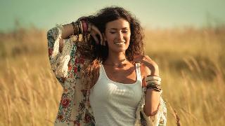 A woman walking through a field of reed grass, eyes closed, smiling, brushing her hair back with her hands.