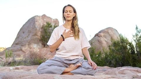 A woman sitting outside in a meditation pose, hand over her heart.
