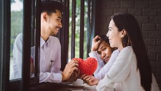 A couple holding a plush red heart between them, symbolizing their love, a man seen in the background looking at them with jealousy.