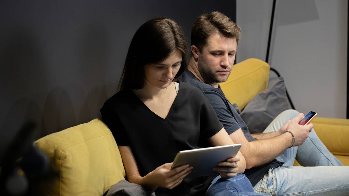 A couple sitting next to each other on the couch, the man looking over his shoulder at the iPad the woman is holding.