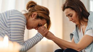 A mother and daughter sitting across from one another, the mother taking her daughter's hands in her own and bending down to press her forehead to them. They both look sad and pained.