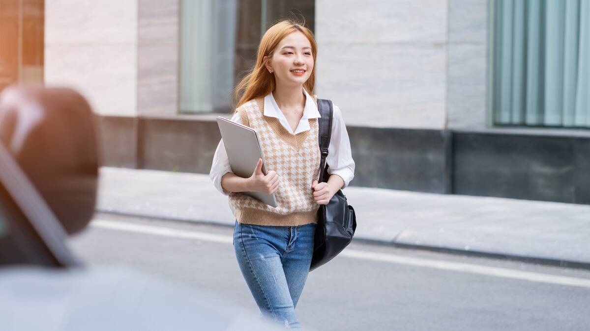 A happy, confident woman holding a laptop in one arm, a shoulder bag in the other, walking to work.