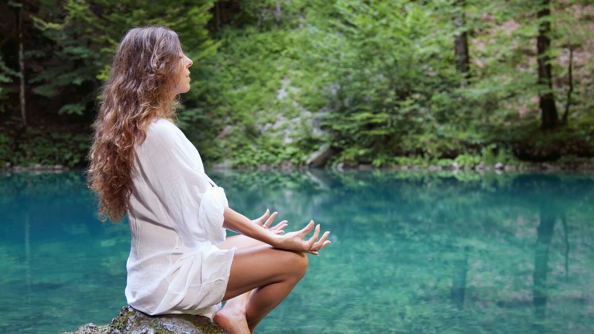 A woman sitting on a rock by the side of a creek, meditating with her eyes closed.