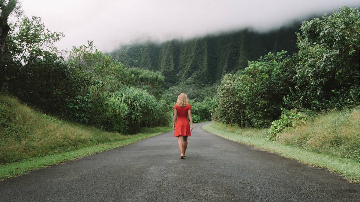 A woman in a bright red dress walking down the center of a paved road that's surrounded by trees, bushes, and fog.
