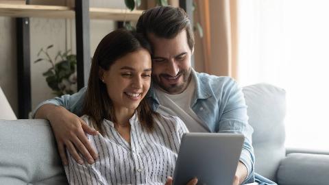 A couple sitting side by side, the man's arm around the woman, both smiling as they look at a tablet screen together.