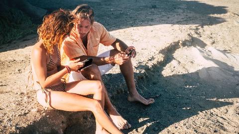 A couple sitting on the beach, the woman showing the man something on her phone.