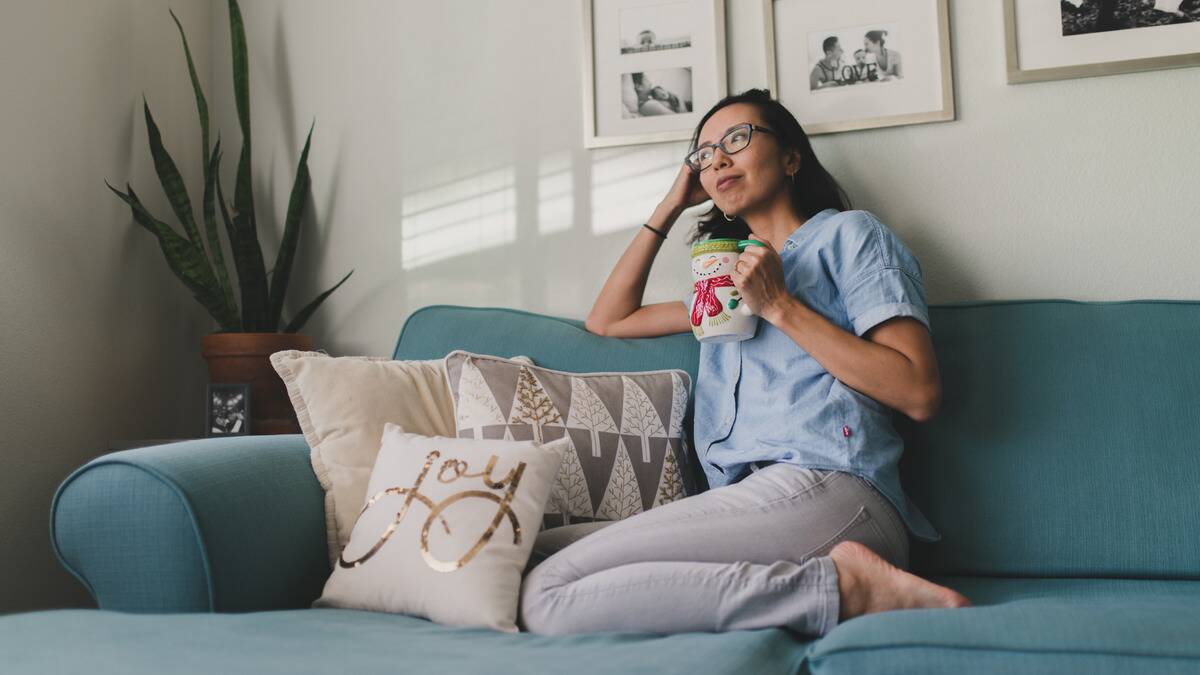 A woman sitting on her couch at home, leaning her head in one hand, holding a mug in the other.