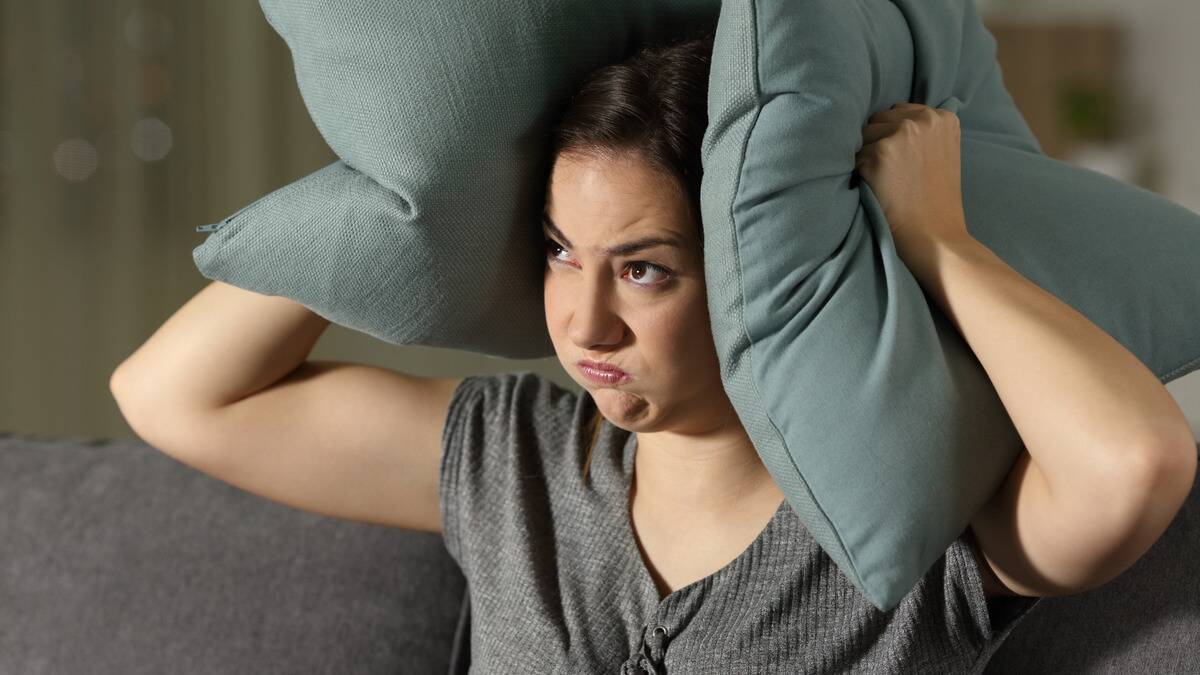 A woman appearing angry, holding up two pillows to her ears to block out noise.