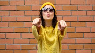 A woman in a yellow hat, yellow sweater, and sunglasses pointing at the camera with both hands, standing in front of a brick wall.