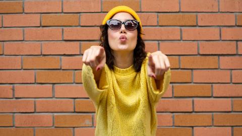 A woman in a yellow hat, yellow sweater, and sunglasses pointing at the camera with both hands, standing in front of a brick wall.