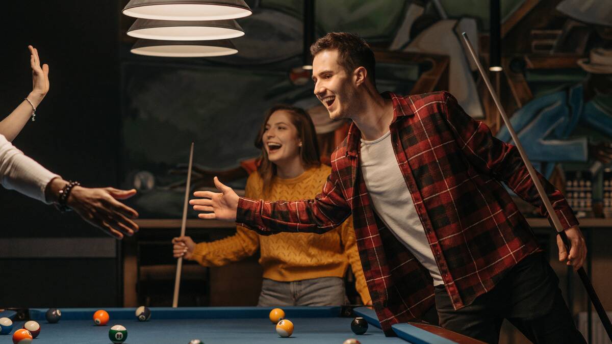 Friends cheering while playing billiards, one reaching across the table to high five someone out of frame.