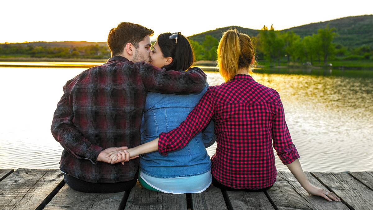 Three people sitting side-by-side on a dock, one man then two women. The man is embracing and kissing the woman directly next to him, but behind her back, he's holding the second woman's hand as well.