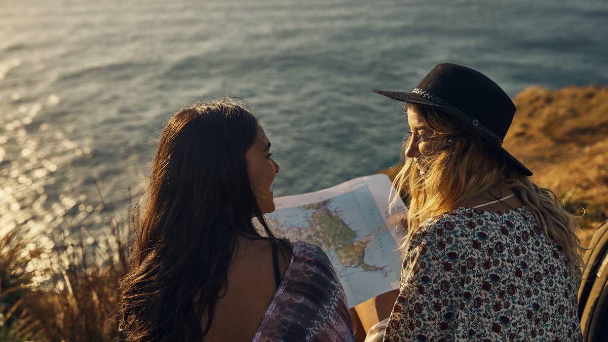 A photo from behind of two friends sat by the water, a map open between them, smiling at one another.