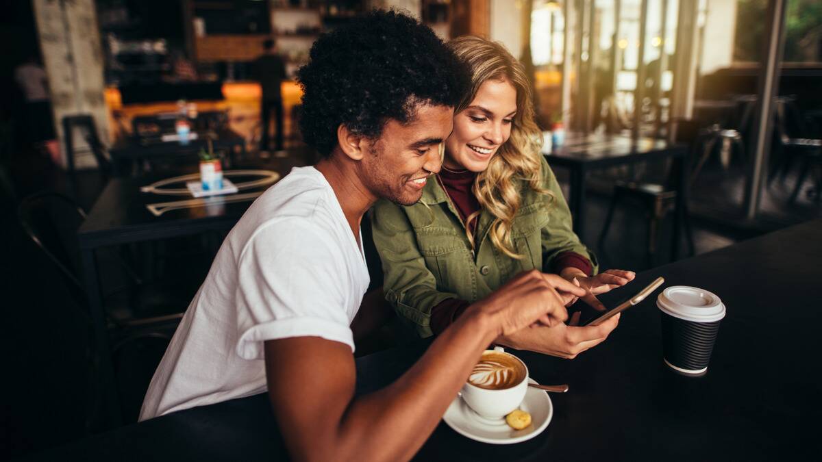 Two friends sat side by side at a cafe, both smiling as they look and point at one of the friends' phones.