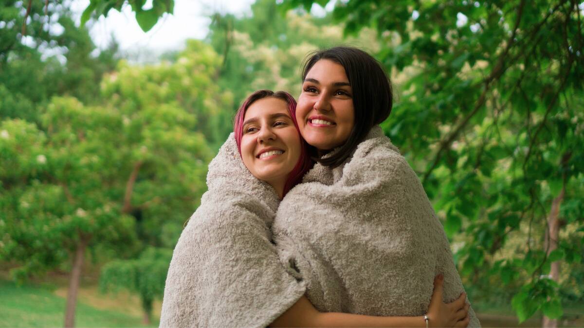 Two friends standing outside, both wrapped in a large blanket, hugging each other.