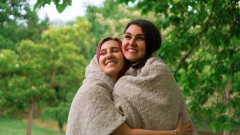 Two friends standing outside, both wrapped in a large blanket, hugging each other.