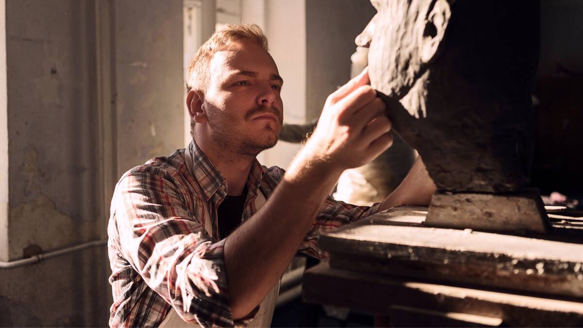A man kneeling down, adding clay to a large sculpture.